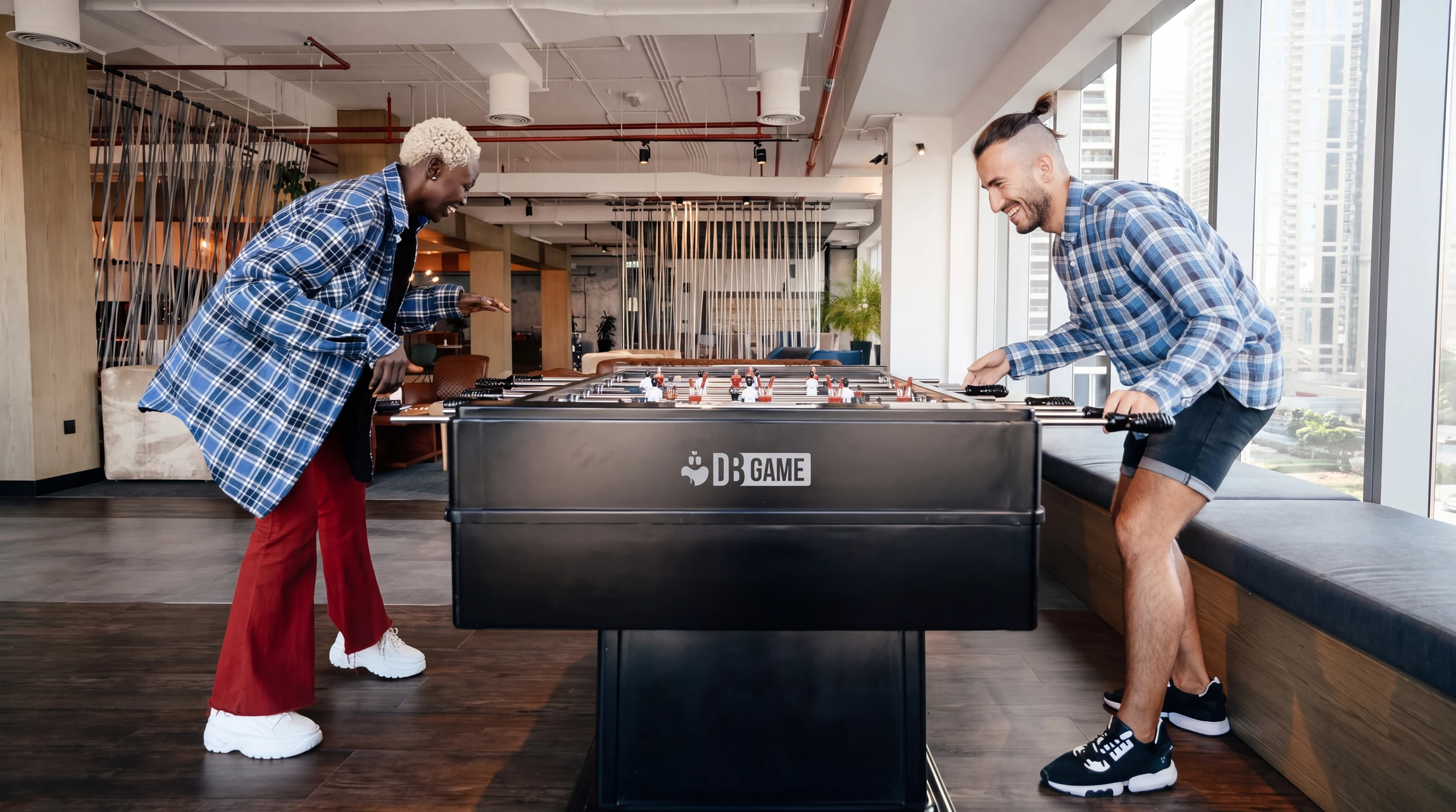 Deux hommes souriants jouent au baby-foot dans un espace de détente moderne, lumineux, avec vue urbaine